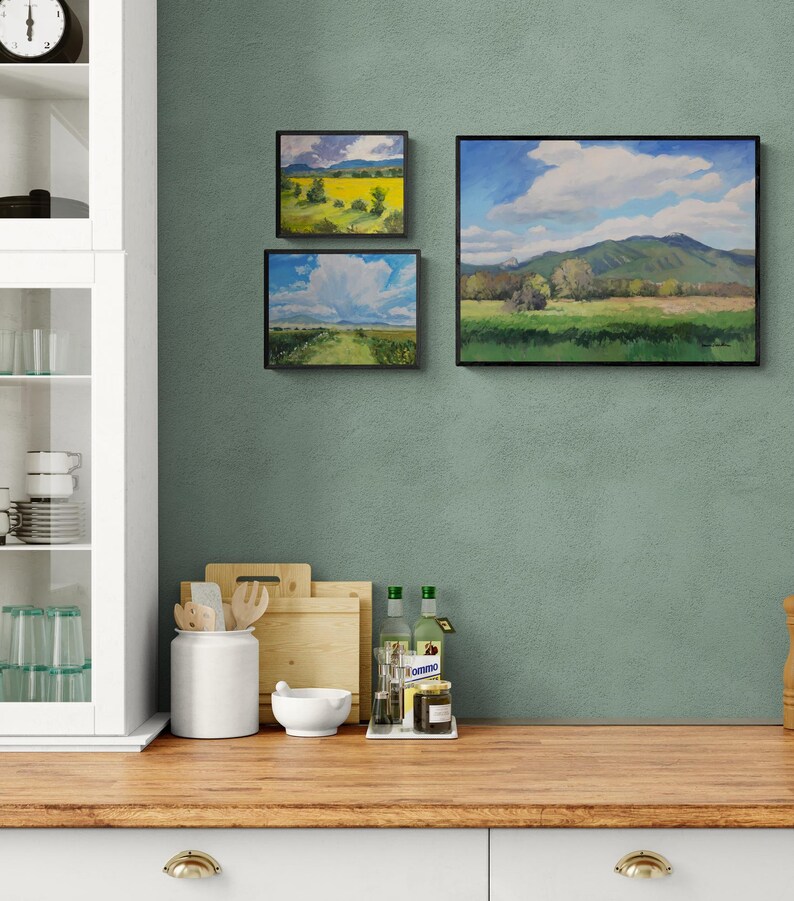 Original New Mexico landscape oil paintings by Dawn Chandler, including Abiquiu Field of Summer Gold, arranged as a small gallery wall in a sage green kitchen above a wooden countertop with cutting boards, glassware, and olive oil bottles, showing how her bright Southwest skies and fields add color to everyday spaces.
