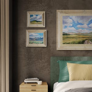 Interior bedroom scene featuring two original New Mexico landscape paintings by Santa Fe artist Dawn Chandler, including Spring Morning Over the Taos Mountains, each framed in light wood and hung on a rich brown wall beside a larger coordinating landscape, demonstrating how the vivid skies, mountains, and open fields bring peaceful Southwestern color and a sense of spaciousness above a contemporary bed and nightstand.