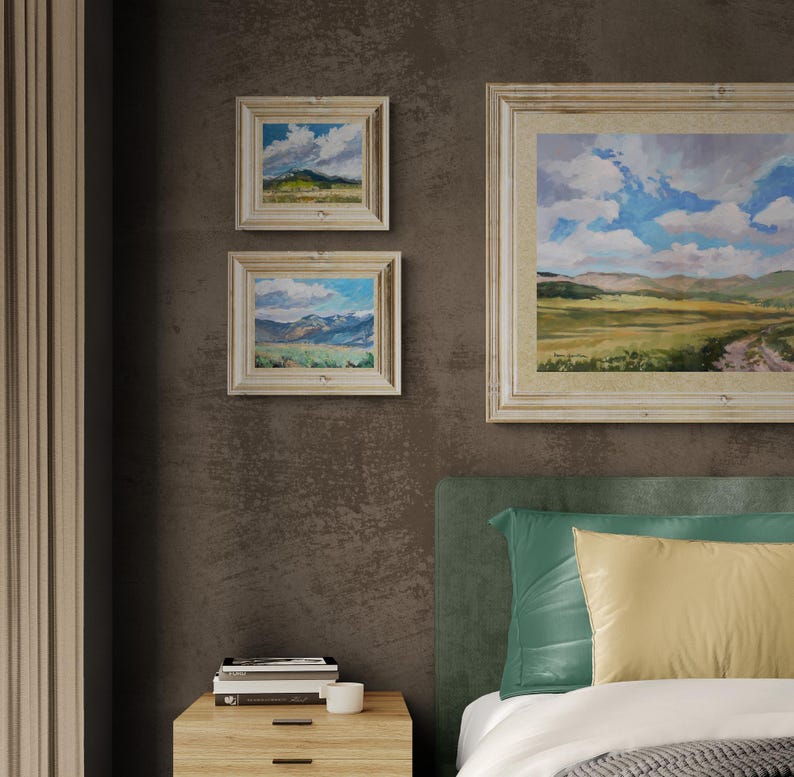 Interior bedroom scene featuring two original New Mexico landscape paintings by Santa Fe artist Dawn Chandler, including Spring Morning Over the Taos Mountains, each framed in light wood and hung on a rich brown wall beside a larger coordinating landscape, demonstrating how the vivid skies, mountains, and open fields bring peaceful Southwestern color and a sense of spaciousness above a contemporary bed and nightstand.