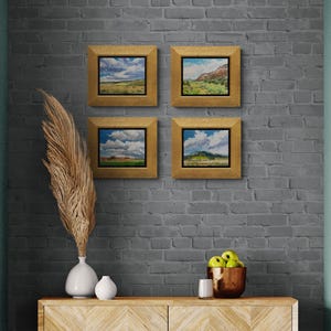 Group of four original New Mexico landscape oil paintings by Santa Fe artist Dawn Chandler, including “Finding My Way to Pedernal,” displayed in gold frames on a gray brick wall above a contemporary console with vases, pampas grass, green apples and candle, showing how these colorful Southwest sky and mesa scenes create a cohesive gallery wall.