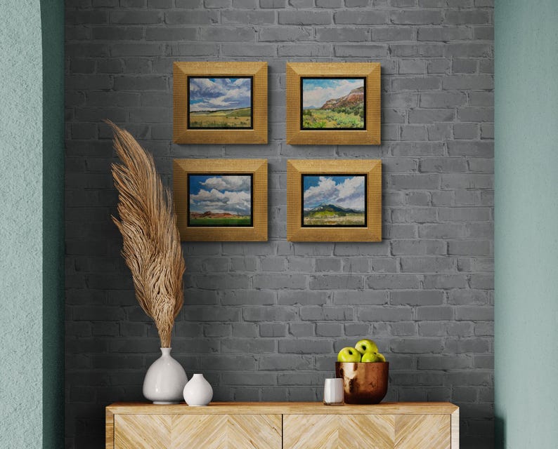Group of four original New Mexico landscape oil paintings by Santa Fe artist Dawn Chandler, including “Finding My Way to Pedernal,” displayed in gold frames on a gray brick wall above a contemporary console with vases, pampas grass, green apples and candle, showing how these colorful Southwest sky and mesa scenes create a cohesive gallery wall.
