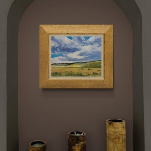 Original New Mexico landscape oil painting Under a High Country August Sky by Santa Fe artist Dawn Chandler displayed in a recessed wall niche, framed in a textured gold frame above three rustic wood candle holders, with the artwork showing expansive golden meadows, rolling green hills, and dramatic August clouds, demonstrating how her high‑country sky painting becomes a focal point in a contemporary Southwestern interior.