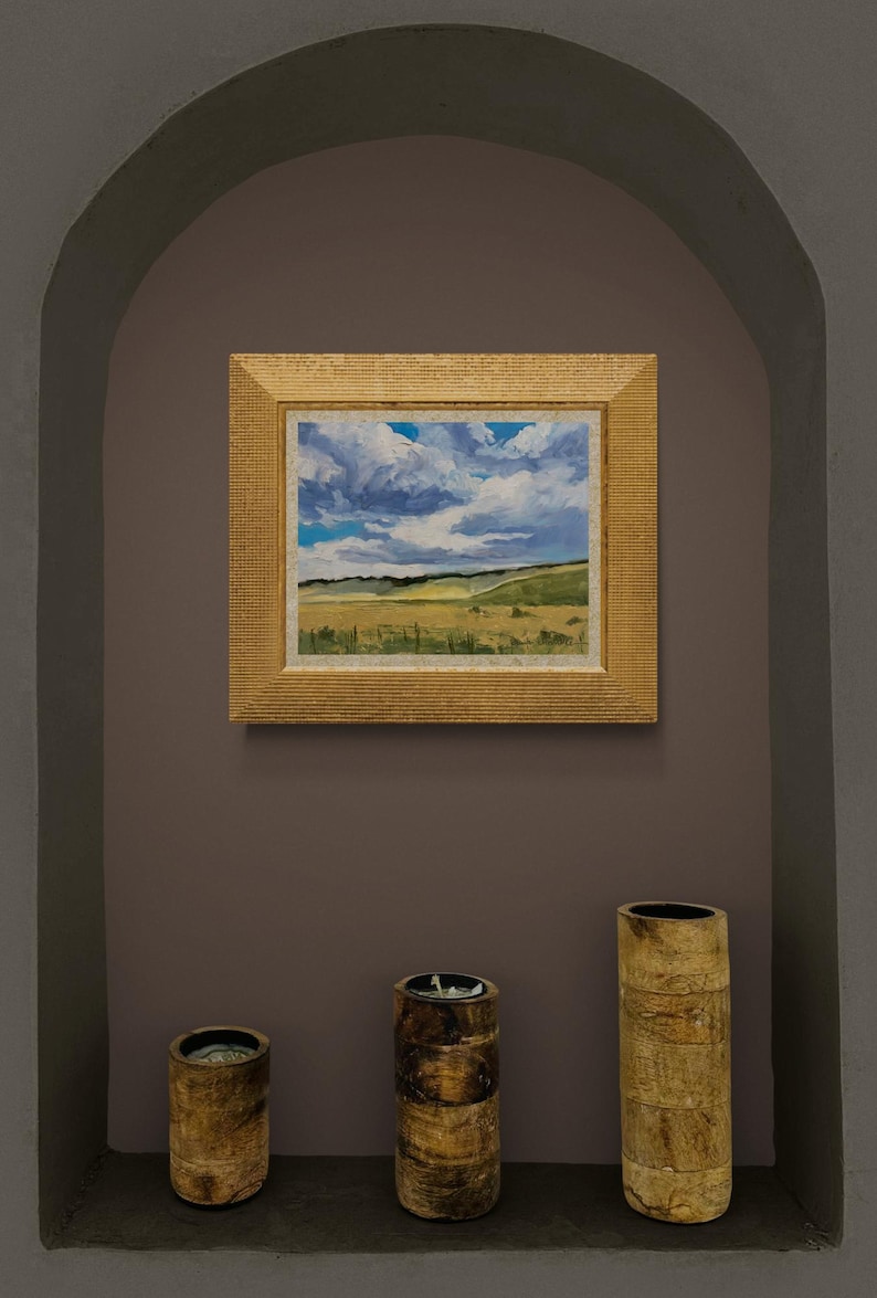 Original New Mexico landscape oil painting Under a High Country August Sky by Santa Fe artist Dawn Chandler displayed in a recessed wall niche, framed in a textured gold frame above three rustic wood candle holders, with the artwork showing expansive golden meadows, rolling green hills, and dramatic August clouds, demonstrating how her high‑country sky painting becomes a focal point in a contemporary Southwestern interior.