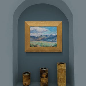 Original New Mexico landscape oil painting Spring Morning Over the Taos Mountains by Santa Fe artist Dawn Chandler shown framed in a warm golden frame and hanging in a recessed blue-gray niche above three simple wooden vessels, illustrating how the colorful Taos valley, blue mountains, and dramatic sky bring vibrant Southwestern mountain art and a sense of spacious calm to a contemporary interior wall.