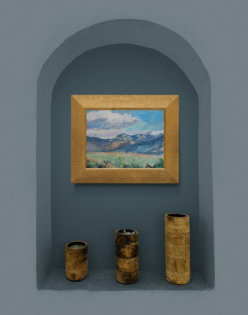 Original New Mexico landscape oil painting Spring Morning Over the Taos Mountains by Santa Fe artist Dawn Chandler shown framed in a warm golden frame and hanging in a recessed blue-gray niche above three simple wooden vessels, illustrating how the colorful Taos valley, blue mountains, and dramatic sky bring vibrant Southwestern mountain art and a sense of spacious calm to a contemporary interior wall.