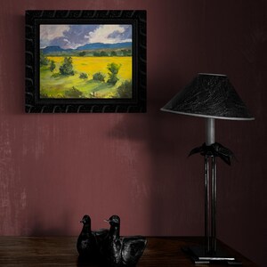 Abiquiu Field of Summer Gold, an original New Mexico landscape oil painting by Santa Fe artist Dawn Chandler, featured in a dark ornate frame on a wine-red wall above a wooden dresser with black sculptural birds and a modern lamp, showing how the vivid field and stormy sky create a dramatic focal point in a bedroom or living room.
