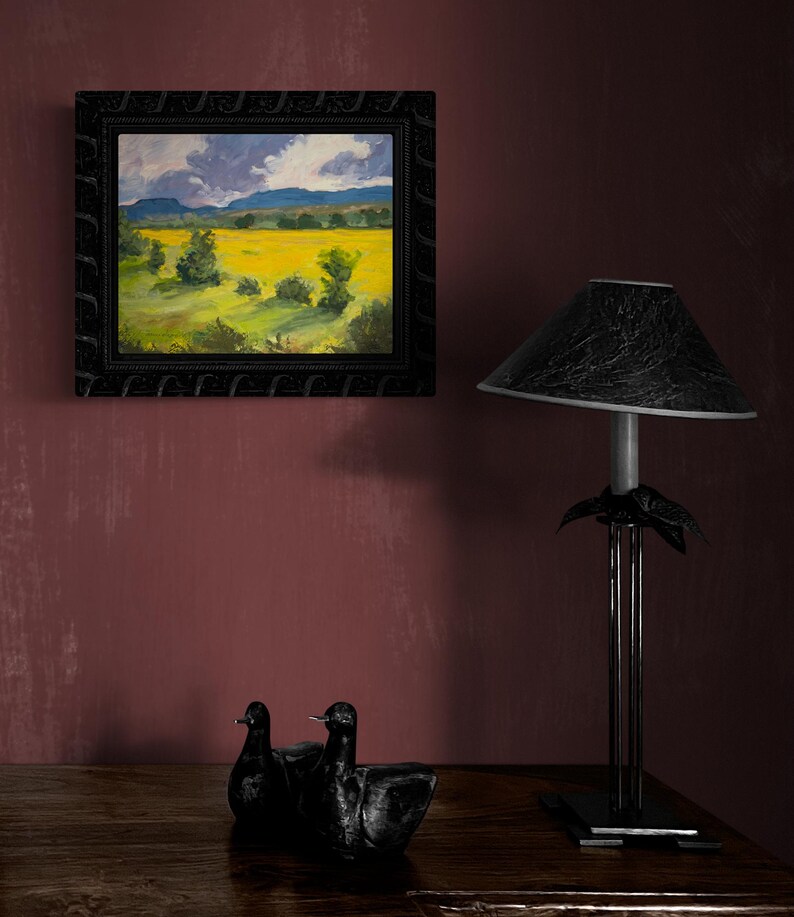Abiquiu Field of Summer Gold, an original New Mexico landscape oil painting by Santa Fe artist Dawn Chandler, featured in a dark ornate frame on a wine-red wall above a wooden dresser with black sculptural birds and a modern lamp, showing how the vivid field and stormy sky create a dramatic focal point in a bedroom or living room.