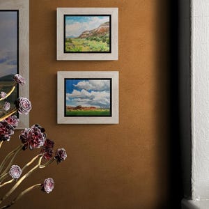 Pair of original New Mexico landscape oil paintings by Santa Fe artist Dawn Chandler, with “Finding My Way to Pedernal” in a light wood frame above another mesa-and-cloudscape, hung on a warm adobe-colored wall beside a window and sculptural succulents, highlighting how these Southwestern skies and valleys bring color and depth to an entry or hallway.