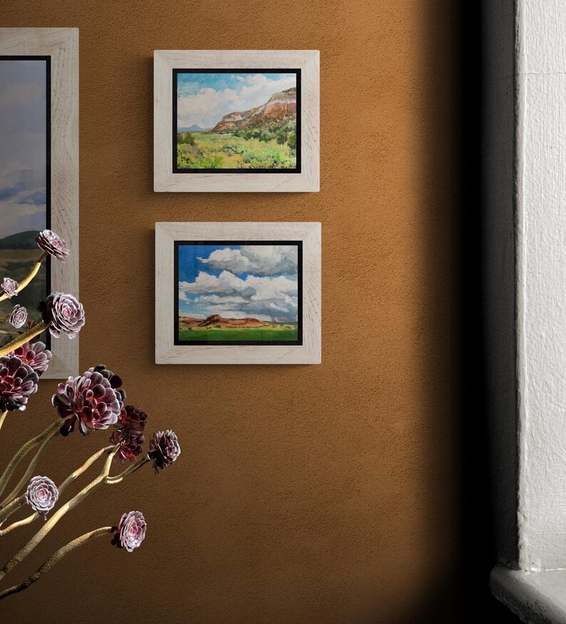 Pair of original New Mexico landscape oil paintings by Santa Fe artist Dawn Chandler, with “Finding My Way to Pedernal” in a light wood frame above another mesa-and-cloudscape, hung on a warm adobe-colored wall beside a window and sculptural succulents, highlighting how these Southwestern skies and valleys bring color and depth to an entry or hallway.