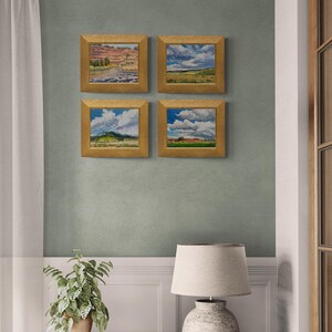 Interior wall scene featuring four framed original New Mexico landscape oil paintings by Santa Fe artist Dawn Chandler, including Under a High Country August Sky and three coordinating high‑country vistas, all in warm golden frames arranged in a grid above a white wainscoted wall, next to a potted plant and neutral lamp, showing how her Southwestern sky and meadow artworks create a cohesive gallery wall in a serene living space.