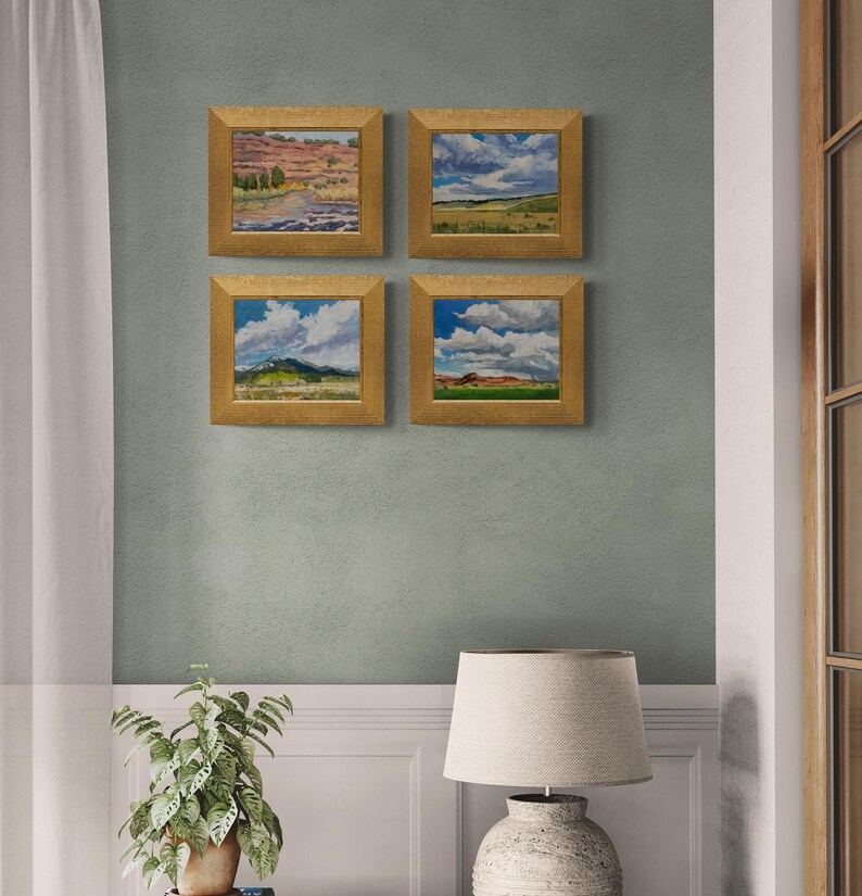 Interior wall scene featuring four framed original New Mexico landscape oil paintings by Santa Fe artist Dawn Chandler, including Under a High Country August Sky and three coordinating high‑country vistas, all in warm golden frames arranged in a grid above a white wainscoted wall, next to a potted plant and neutral lamp, showing how her Southwestern sky and meadow artworks create a cohesive gallery wall in a serene living space.