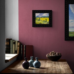 Abiquiu Field of Summer Gold, an original New Mexico landscape oil painting by Santa Fe artist Dawn Chandler, displayed in a black ornate frame on a plum-colored wall above a rustic wooden table with books, small ceramic vases, and a bowl of seed pods, illustrating how the sunny field scene warms an intimate reading nook.