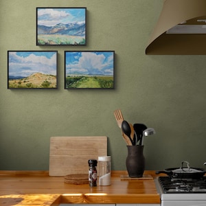 Cozy kitchen interior featuring three original New Mexico landscape paintings by Santa Fe artist Dawn Chandler, including Spring Morning Over the Taos Mountains, framed in simple black frames and arranged on a sage green wall above a warm wood countertop with cooking utensils and spices, illustrating how the bright skies, mesas, and open fields create inviting Southwestern art for everyday living spaces.