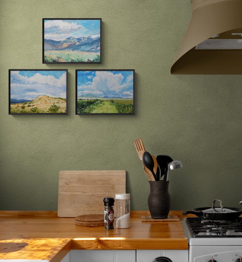 Cozy kitchen interior featuring three original New Mexico landscape paintings by Santa Fe artist Dawn Chandler, including Spring Morning Over the Taos Mountains, framed in simple black frames and arranged on a sage green wall above a warm wood countertop with cooking utensils and spices, illustrating how the bright skies, mesas, and open fields create inviting Southwestern art for everyday living spaces.