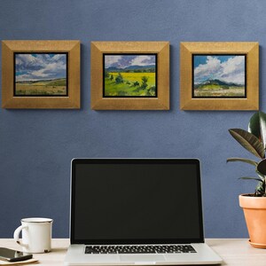 Home office with a laptop, mug, phone, and potted rubber plant on a light wood desk beneath three framed original New Mexico landscape paintings by Santa Fe artist Dawn Chandler, including Under a High Country August Sky at left and two complementary high‑country vistas, all in gold frames against a blue wall, illustrating how her Southwestern sky and field paintings bring color, nature, and calm focus to a creative workspace.