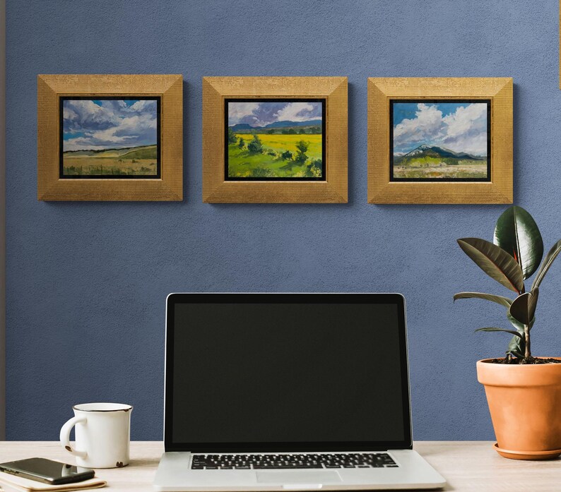 Home office with a laptop, mug, phone, and potted rubber plant on a light wood desk beneath three framed original New Mexico landscape paintings by Santa Fe artist Dawn Chandler, including Under a High Country August Sky at left and two complementary high‑country vistas, all in gold frames against a blue wall, illustrating how her Southwestern sky and field paintings bring color, nature, and calm focus to a creative workspace.