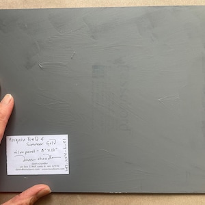 Back view of the original New Mexico landscape oil painting Abiquiu Field of Summer Gold by Santa Fe artist Dawn Chandler, showing the gray-primed 8 x 10 inch panel resting on a tabletop, with Dawn’s hand holding one corner and a neatly printed label indicating the title, medium, size, artist signature, mailing address, email, and website, emphasizing the artwork’s authenticity and professional presentation for collectors.