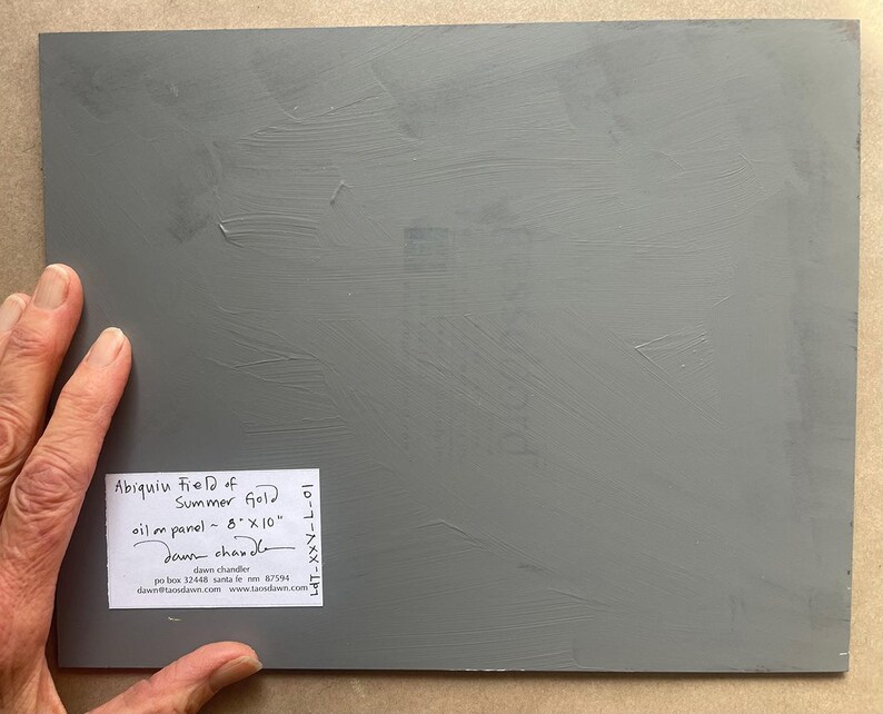 Back view of the original New Mexico landscape oil painting Abiquiu Field of Summer Gold by Santa Fe artist Dawn Chandler, showing the gray-primed 8 x 10 inch panel resting on a tabletop, with Dawn’s hand holding one corner and a neatly printed label indicating the title, medium, size, artist signature, mailing address, email, and website, emphasizing the artwork’s authenticity and professional presentation for collectors.