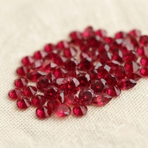 May include: A collection of numerous small, round, faceted red gemstones. Each stone features a brilliant cut with multiple reflective surfaces that catch the light, creating a deep crimson color profile across the entire cluster of loose gems.