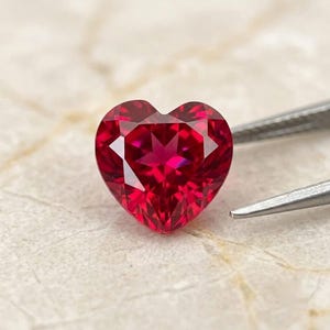 May include: A heart-shaped, faceted gemstone with a deep, vibrant red color. The stone features precise geometric cuts that create a star-like pattern of light reflection across its surface, held by a pair of thin, silver-toned metal tweezers.
