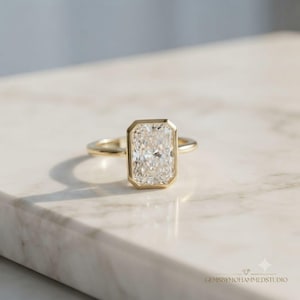 May include: A gold ring featuring a rectangular radiant-cut centre stone secured in a bezel setting. The band is smooth and polished gold. The text GEMSBYMOHAMMEDSTUDIO is visible in the bottom right corner of the image.