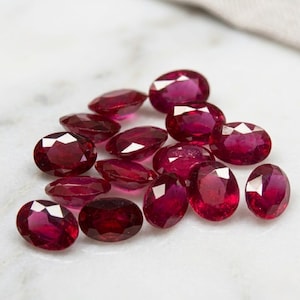 May include: A collection of loose, oval-cut rubies featuring a deep, vibrant red color and faceted surfaces that reflect light. These gemstones are arranged in a cluster, showcasing their polished, translucent appearance and uniform shape.