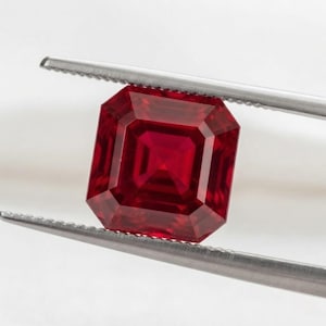 Può includere: Una gemma rosso vivo e profondo con un taglio a gradini quadrato, tenuta saldamente tra le punte metalliche seghettate di una pinzetta color argento. La pietra mostra una ricca tonalità cremisi satura con chiari riflessi geometrici interni.