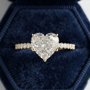 Op de afbeelding: Een gouden ring met een grote, hartvormige diamant in het midden, vastgehouden door vier pootjes. De ringband is dun en versierd met een rij kleine, rondgeslepen diamanten die zich aan beide kanten van de centrale steen uitstrekken.
