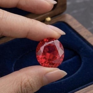 May include: A round, faceted, vibrant red gemstone with a brilliant cut, displaying deep crimson hues and sharp geometric facets that reflect light across its surface, held between fingers above a dark wooden box with a blue velvet interior.