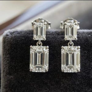 Puede incluir: Un par de pendientes colgantes con dos piedras transparentes de corte esmeralda en metal plateado. Cada pendiente consta de una piedra rectangular pequeña en la parte superior conectada a una piedra rectangular más grande debajo, con cierre de presión.