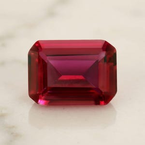 Può includere: Una gemma di un rosso profondo e vibrante, tagliata in una forma rettangolare a smeraldo. La pietra presenta bordi sfaccettati precisi che creano un chiaro motivo geometrico, con la luce che si riflette attraverso il corpo cremisi traslucido.