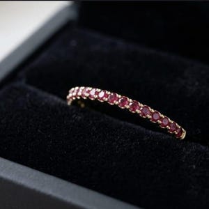 Könnte beinhalten: Ein dünner Goldring mit einer durchgehenden Reihe kleiner, runder, tiefroter Rubine, die in der oberen Hälfte gefasst sind. Der Ring liegt in einer mit schwarzem Samt ausgekleideten Schmuckschatulle, was den Kontrast der Materialien betont.