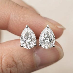 May include: A pair of stud earrings featuring clear, pear-shaped faceted stones set in a three-prong silver-toned metal setting. The stones are transparent and reflect light with a brilliant, multi-faceted appearance, secured by a simple metal frame.