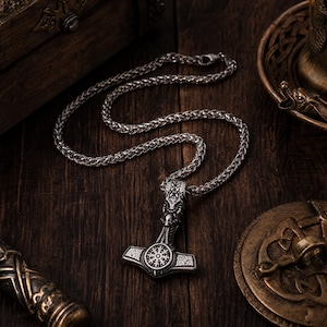May include: A silver-coloured metal pendant shaped like a hammer, featuring intricate engraved patterns and a circular symbol with radiating lines at its centre, suspended from a matching silver-coloured woven chain necklace.