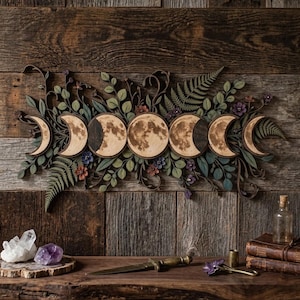 Puede incluir: Un adorno de pared horizontal de madera que muestra las fases lunares desde la luna creciente hasta la luna llena y viceversa, rodeado de follaje intrincado cortado con láser, helechos y pequeñas flores en tonos verdes, marrones y morados.