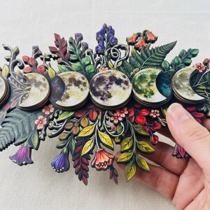 Pu&ograve; includere: Una decorazione da parete con una serie di cinque fasi lunari circolari disposte orizzontalmente. I dischi lunari sono circondati da intricate e colorate illustrazioni di vari fiori, felci e foglie nelle tonalit&agrave; del verde, rosso e viola.