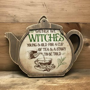 Może przedstawiać: Drewniana tabliczka ścienna w kształcie czajnika z postarzanym wykończeniem. Tekst brzmi: GATHER WE WITCHES YOUNG & OLD FOR A CUP OF TEA & A STORY TO BE TOLD. Zawiera grafikę czajnika nalewającego herbatę do filiżanki oraz małą ilustrację liścia.