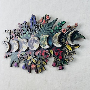 Pu&ograve; includere: Una decorazione da parete con una disposizione orizzontale delle fasi lunari, dalla falce alla luna piena, circondata da illustrazioni dettagliate di felci, foglie e vari fiori colorati nelle tonalit&agrave; del rosso, viola, giallo e verde.