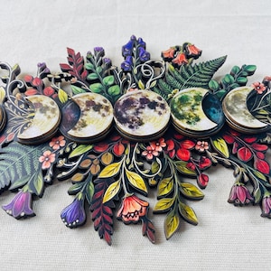 Pu&ograve; includere: Una decorazione da parete con cinque fasi lunari circolari disposte orizzontalmente, sovrapposte a un'illustrazione dettagliata di elementi botanici colorati, tra cui felci, foglie e vari fiori nelle tonalit&agrave; del viola, rosso, giallo e verde.