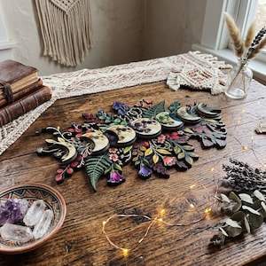 Pu&ograve; includere: Una decorazione da parete in legno con una disposizione orizzontale delle fasi lunari circondate da elementi botanici dipinti dai colori vivaci, tra cui felci verdi, bacche rosse e fiori viola, realizzati con dettagli intagliati al laser.