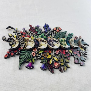 Pu&ograve; includere: Una decorazione da parete con una disposizione orizzontale di sette fasi lunari in vari stadi di illuminazione, circondate da complesse e colorate illustrazioni botaniche di felci, foglie e fiori nelle tonalit&agrave; del viola, rosso e verde.