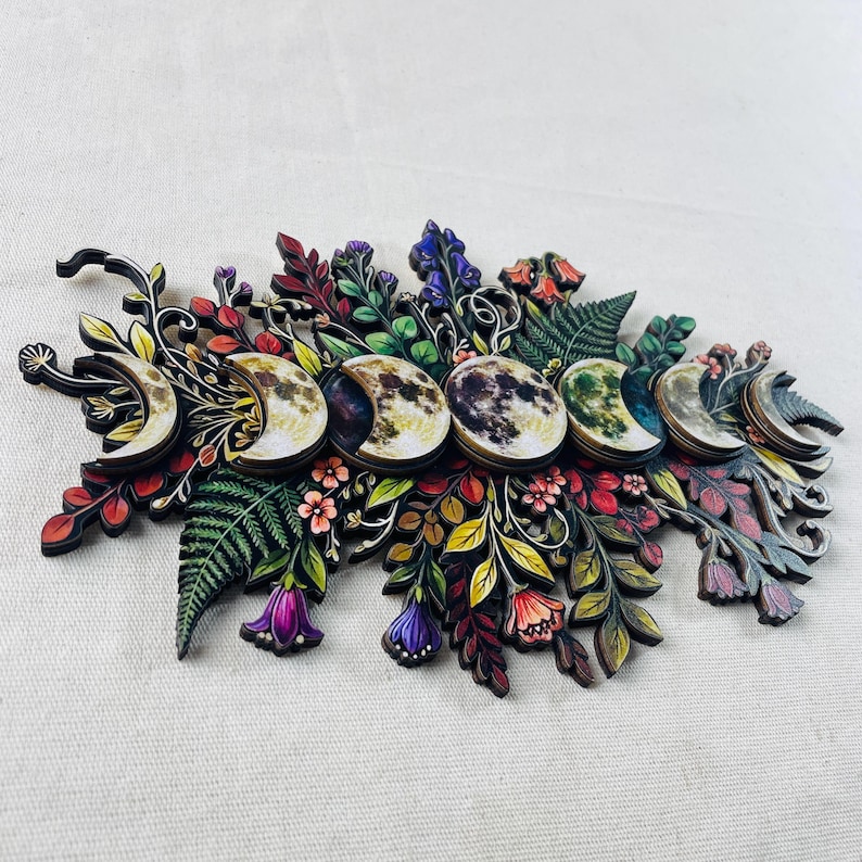 Pu&ograve; includere: Una decorazione da parete con una disposizione orizzontale di sette fasi lunari in vari stadi di illuminazione, circondate da complesse e colorate illustrazioni botaniche di felci, foglie e fiori nelle tonalit&agrave; del viola, rosso e verde.