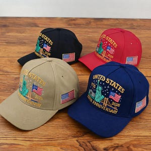 Puede incluir: Cuatro gorras de béisbol en negro, rojo, beige y azul marino. Cada gorra presenta el bordado United States 250th Anniversary 1776-2026, la Estatua de la Libertad, un puente y un pequeño parche de la bandera estadounidense en el lateral.
