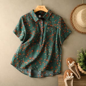 May include: A short-sleeved button-down shirt in a dark teal green fabric featuring a small-scale floral pattern with orange and white blossoms. The shirt has a pointed collar, a single chest pocket on the left side, and a slightly curved hemline.