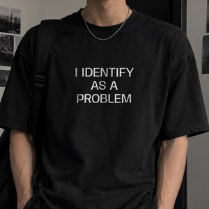Op de afbeelding: Een zwart T-shirt met korte mouwen en de witte tekst I IDENTIFY AS A PROBLEM in een strak schreefloos lettertype op de borst. Het kledingstuk heeft een ronde hals en een relaxte pasvorm, gecombineerd met een zwarte rugzak en een donkere spijkerbroek.