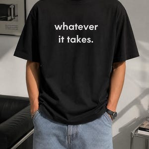 May include: A black short-sleeved t-shirt featuring the white text "whatever it takes." printed in a simple sans-serif font across the chest. The garment has a classic crew neckline and a relaxed fit, paired with light blue denim jeans.