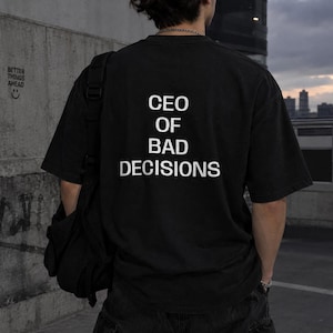 May include: A black short-sleeved t-shirt featuring the white text CEO OF BAD DECISIONS printed on the back. The shirt is worn with a black shoulder bag and dark pants, showcasing a casual style with a bold graphic message on the fabric.