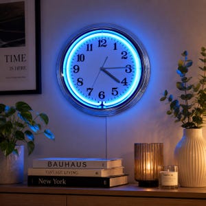 Puede incluir: Un reloj de pared redondo con esfera blanca, números negros y manecillas negras, rodeado por un anillo de luz de neón azul brillante. El reloj cuenta con un marco de metal plateado y está montado en la pared, emitiendo un resplandor vibrante.