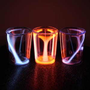 Peut inclure: Trois verres transparents à double paroi. Le verre de gauche présente une lumière interne bleue, le verre central dégage une lueur orange vive et le verre de droite affiche une illumination interne violette douce et diffuse.