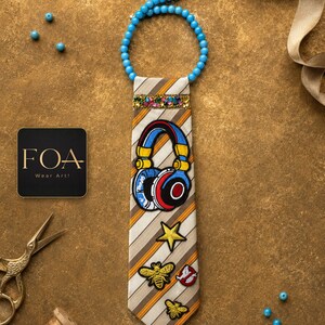 Op de afbeelding: Een beige en bruin gestreepte das met een blauwe kralenhalsband, een band van kleurrijke strass-steentjes en geborduurde patches waaronder een blauwe, rode en gele koptelefoon, een gele ster, twee gouden bijen en een wit spookicoon op een rode cirkel.